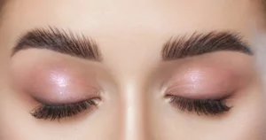 Close-up of closed eyes with thick, dark brows, shimmery pink eyeshadow, and long lashes featuring Brow Extensions in Martinsville, VA.